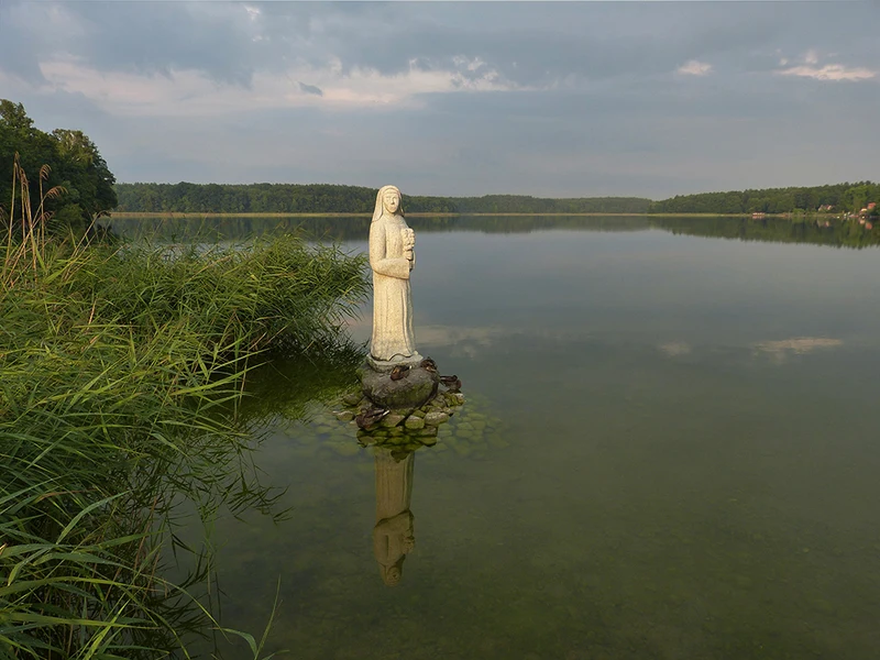 Der Wutzsee bei Lindow mit bewaldetem Ufersaum, im Vordergrund die Skulptur der Schönen Nonne