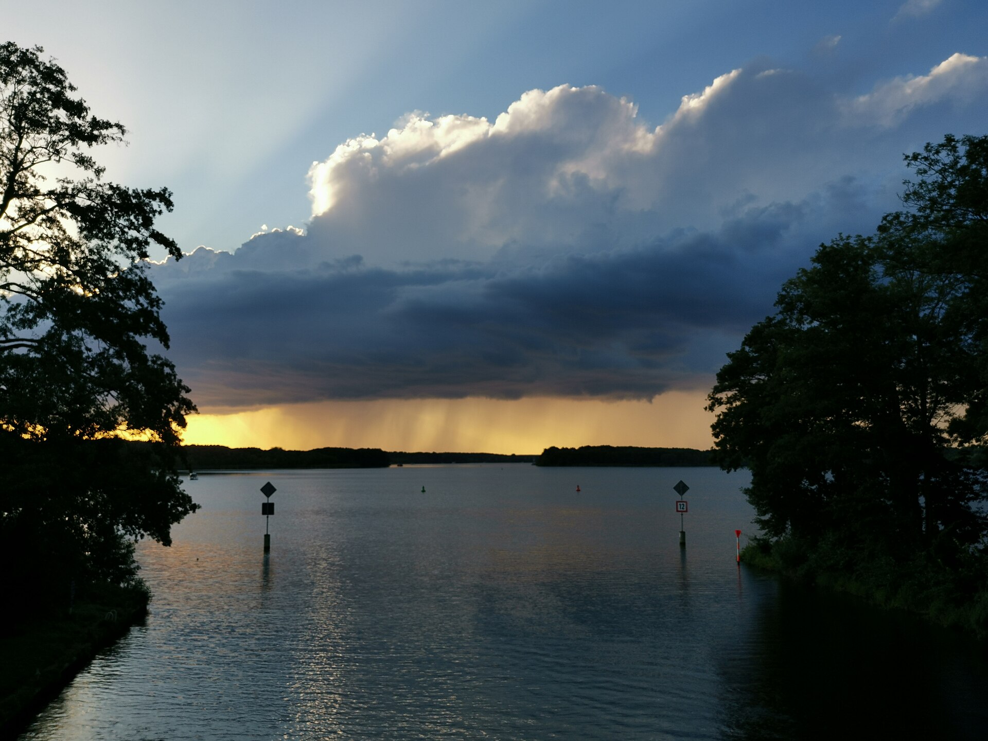 Regenschauer über dem Dolgensee bei Dolgenbrodt, links und rechts Bäume, auf dem Wasser zwei Fahrwasserzeichen